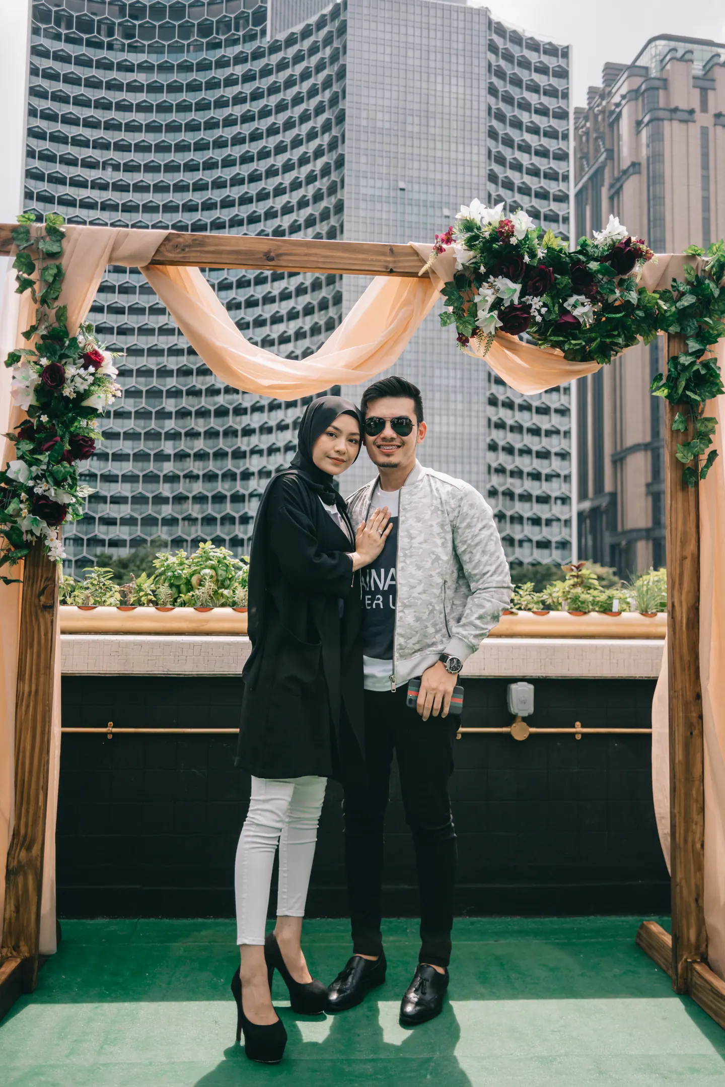 A couple stand under a floral arch with towers behind.
