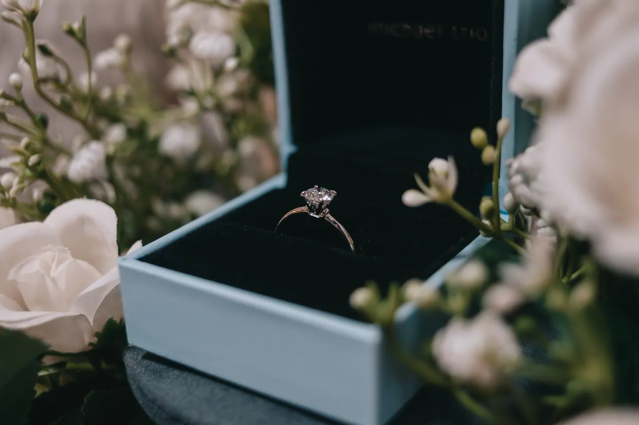 A diamond ring in an open blue box among white flowers.