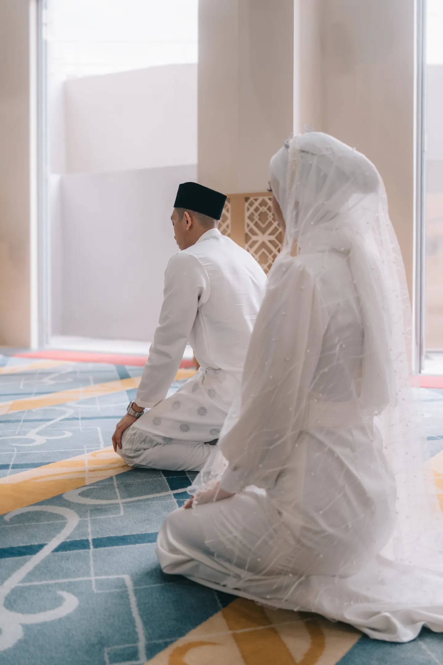 Bride and groom kneel side by side on a patterned carpet, seen from behind.