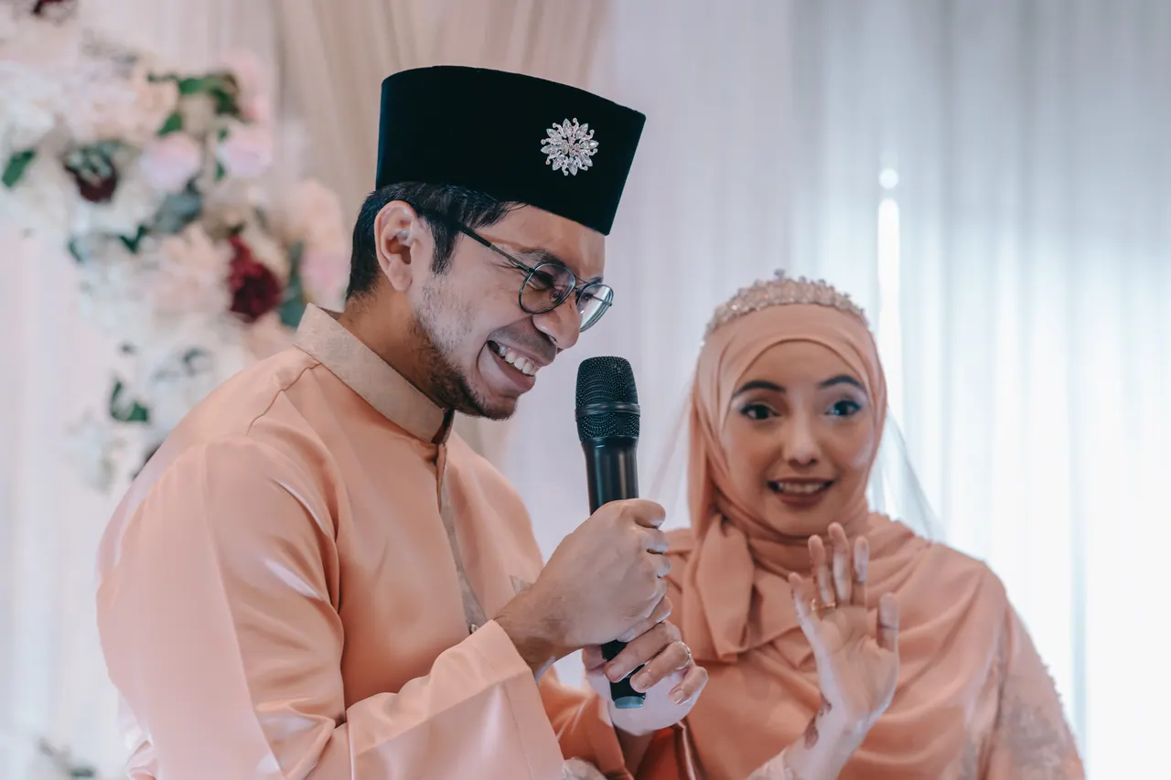 The groom speaks into a microphone while the bride laughs and waves.