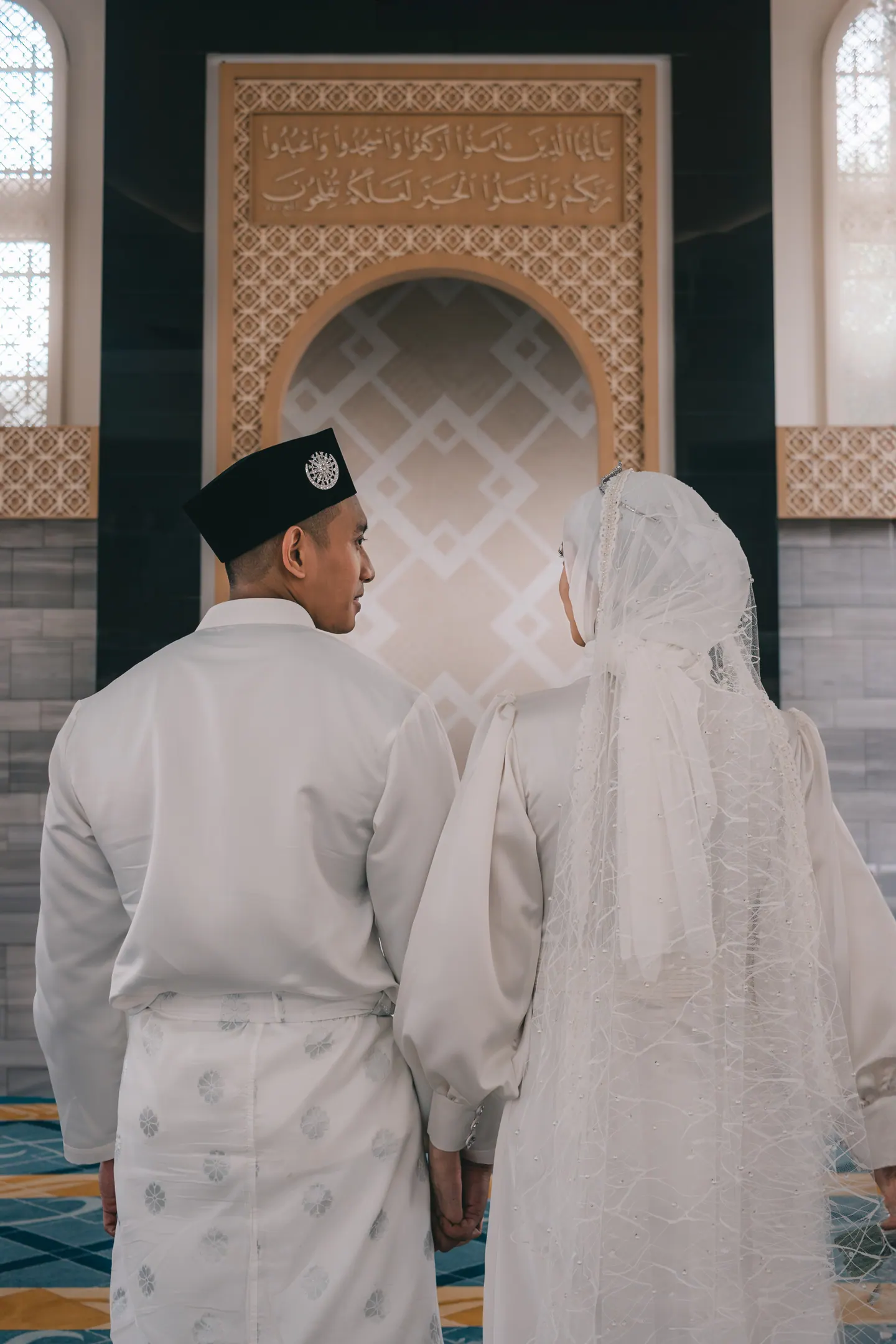 Bride and groom stand hand in hand before a carved niche, seen from behind.