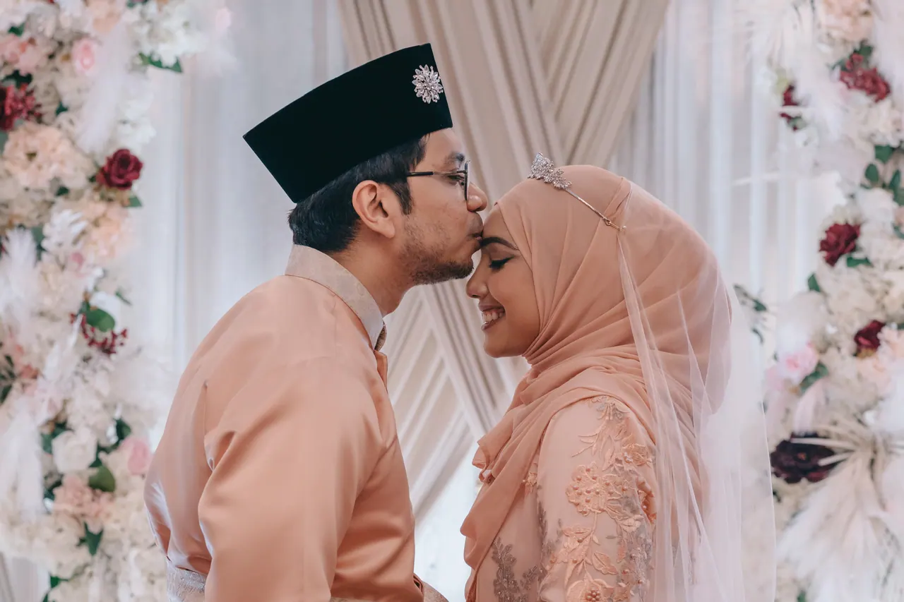The groom kisses the bride’s forehead under the floral arch.