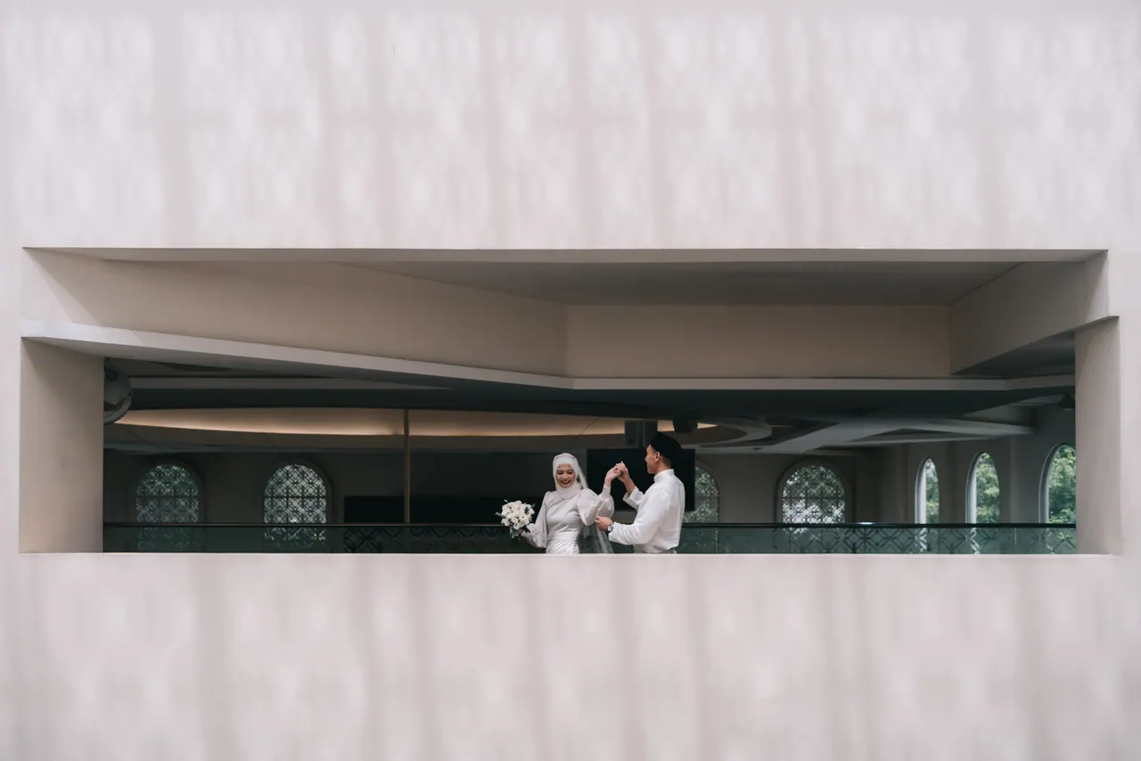 Bride and groom, small inside a long window cut into a pale wall.