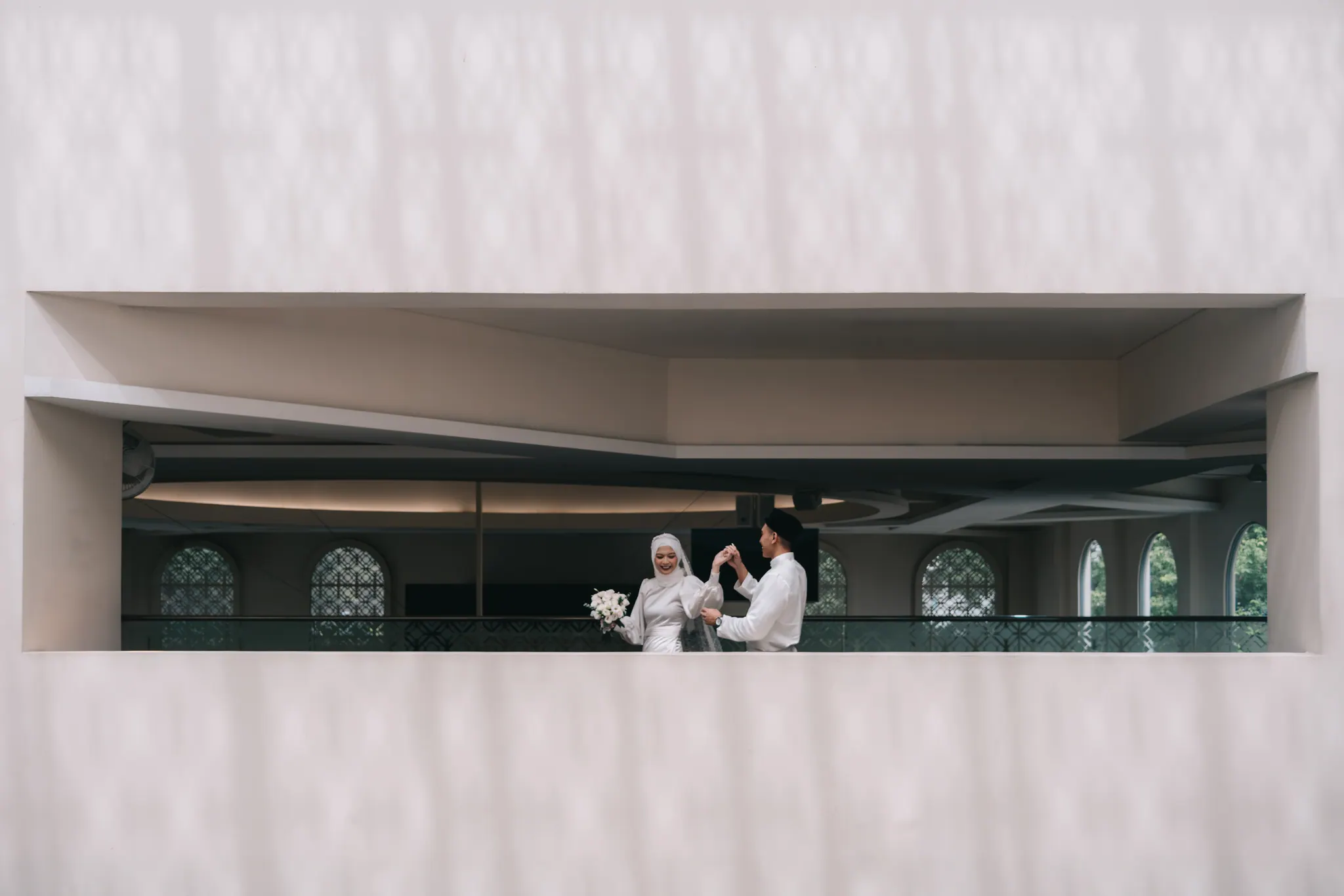 Bride and groom, small inside a long window cut into a pale wall.