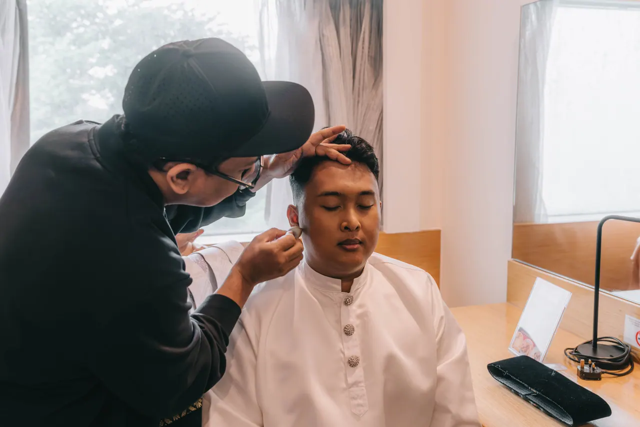A make-up artist powders the groom’s face by the window.