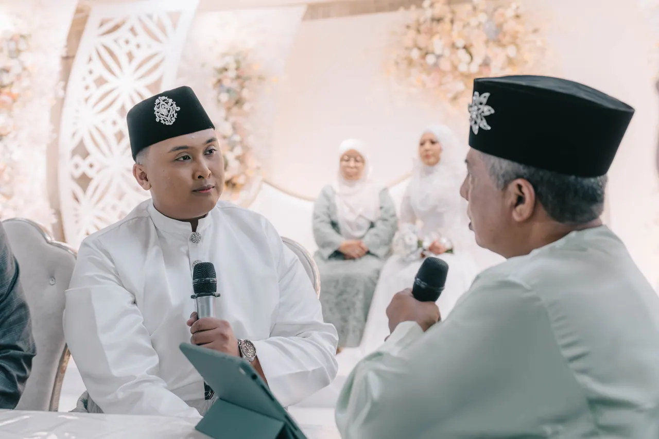 The groom and an older man face each other with microphones at the ceremony table.