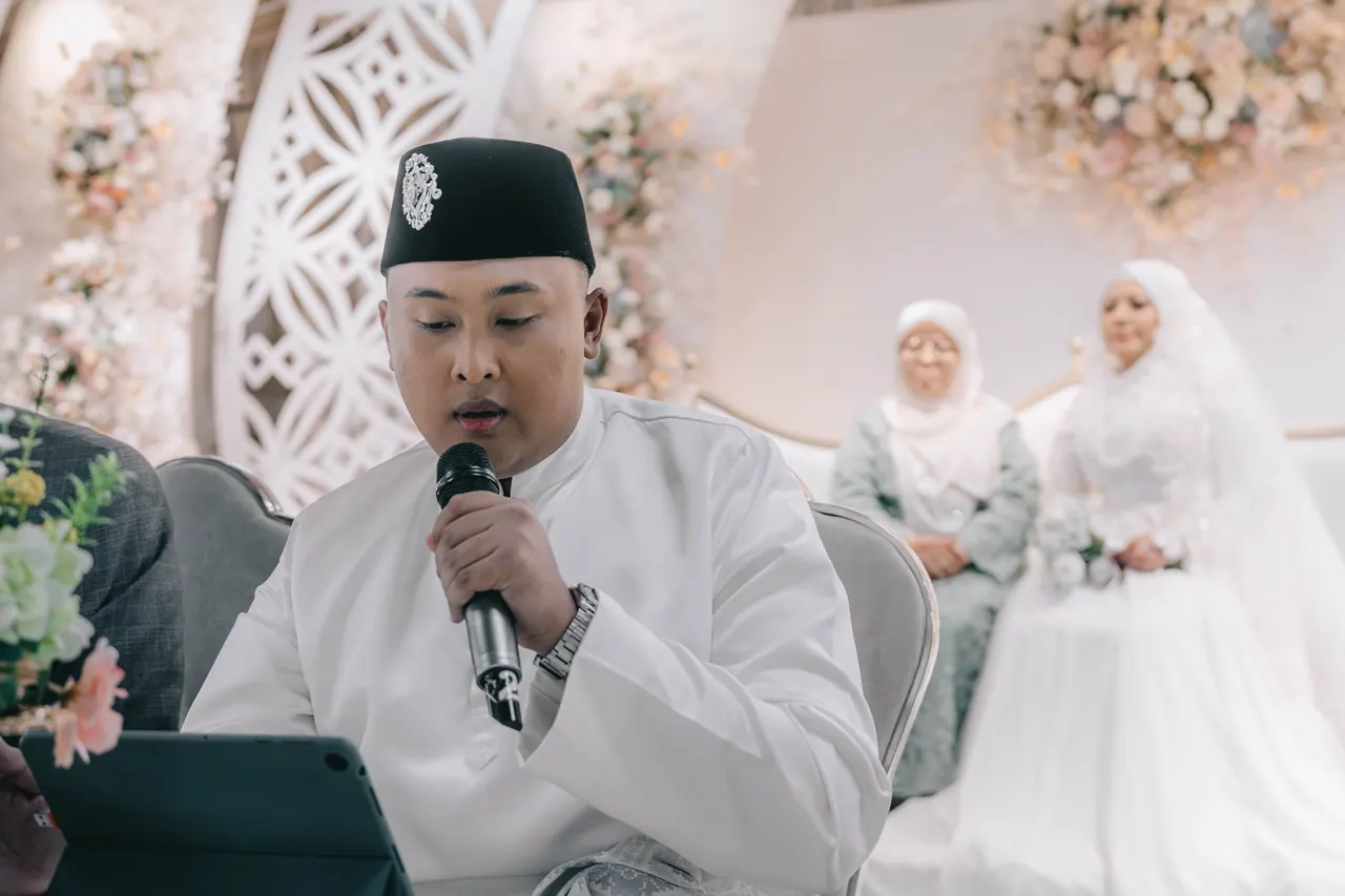 The groom speaks into a microphone, the bride seated behind him.