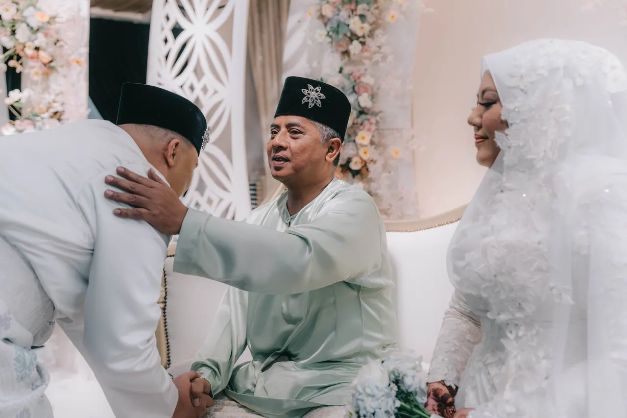 An older man rests a hand on the groom’s shoulder; the bride smiles beside them.