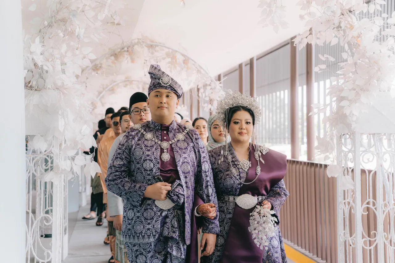 The couple, in purple and silver, lead a procession under a white arch.