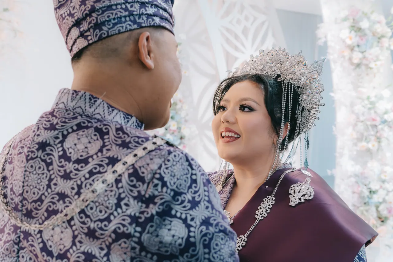 Over the groom’s shoulder, the bride in a silver headdress smiles at him.