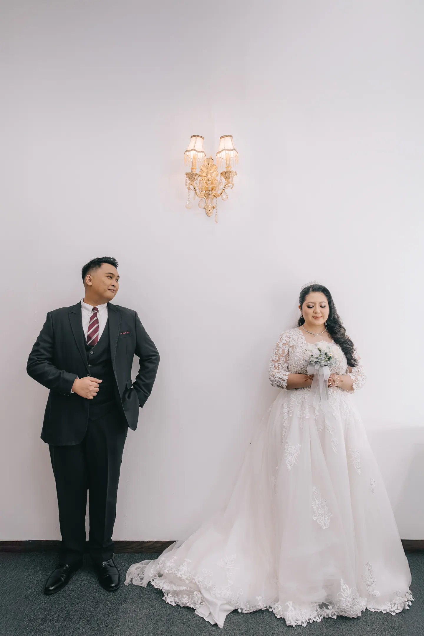 Bride and groom stand apart against a tall white wall beneath a single brass sconce.