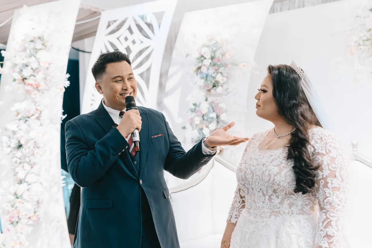 The groom, holding a microphone, gestures towards the bride in her lace gown.
