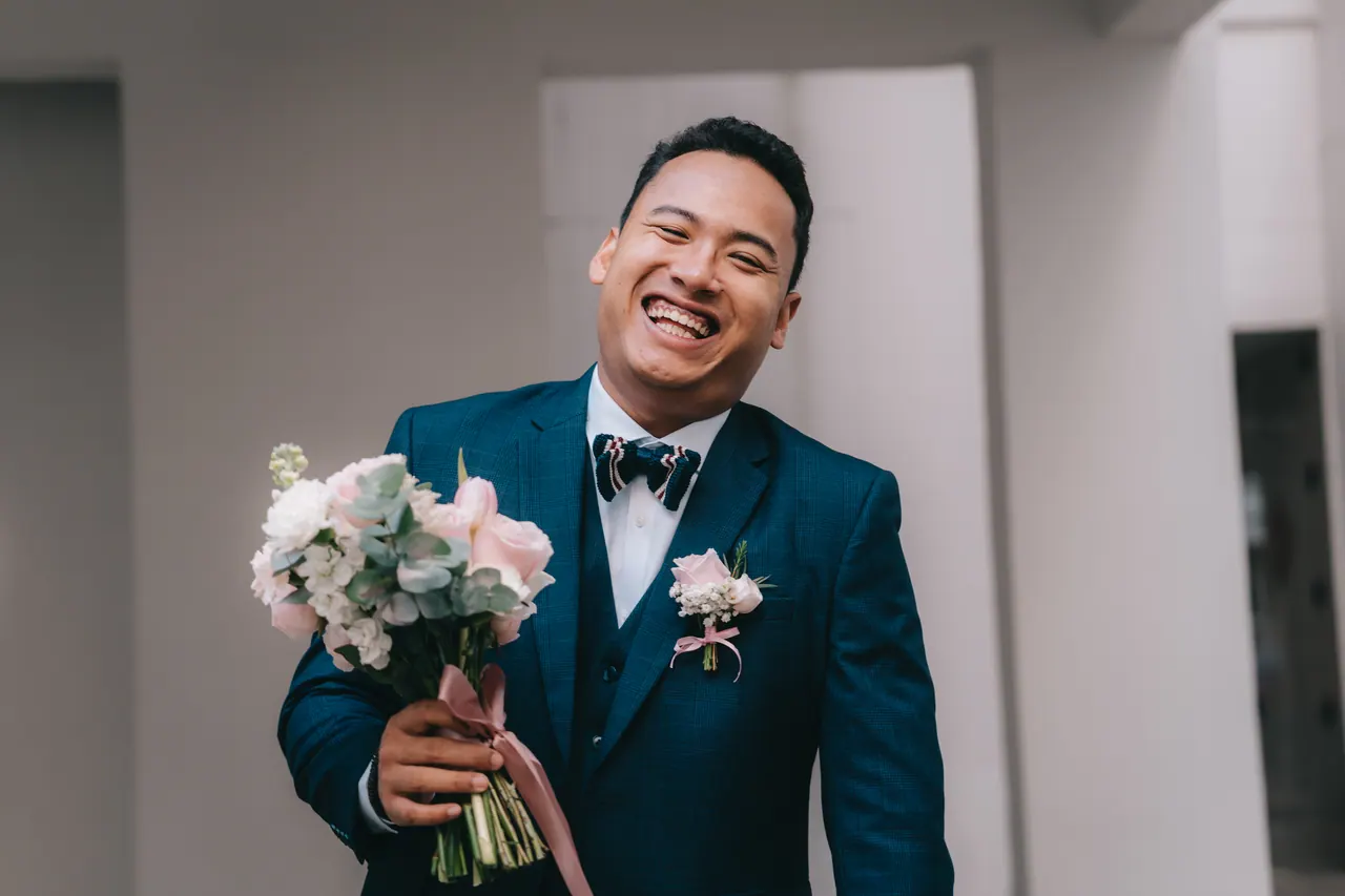 The groom laughs, holding a bouquet of pink roses.