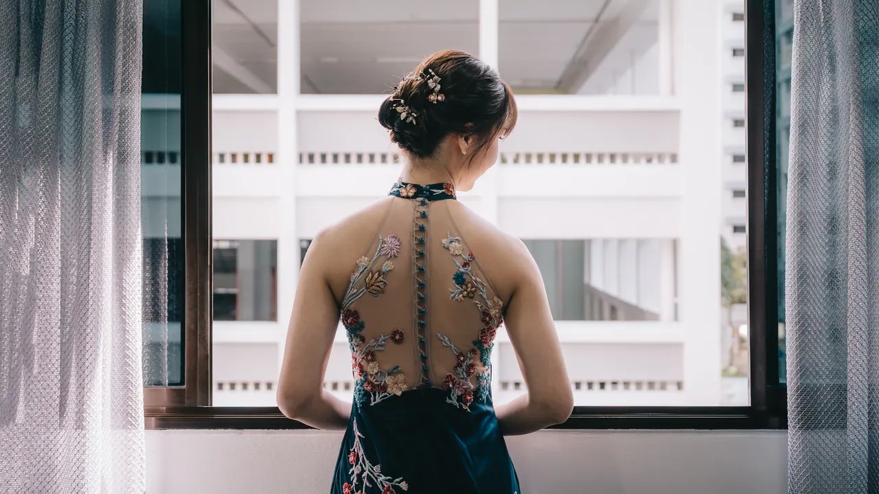 The bride, in an embroidered cheongsam with a sheer back, stands at a window.