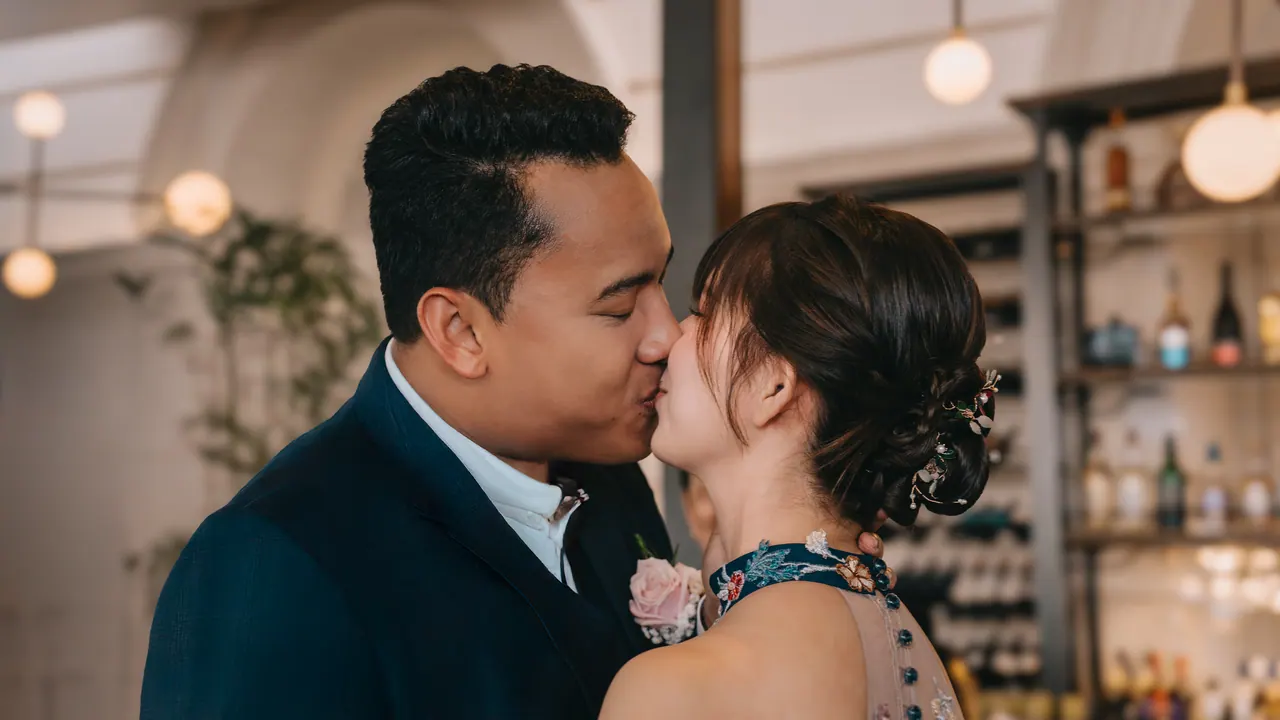 Bride and groom kiss in front of a lit bar.
