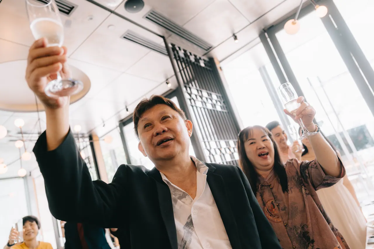 An older man raises his glass high, guests behind him.