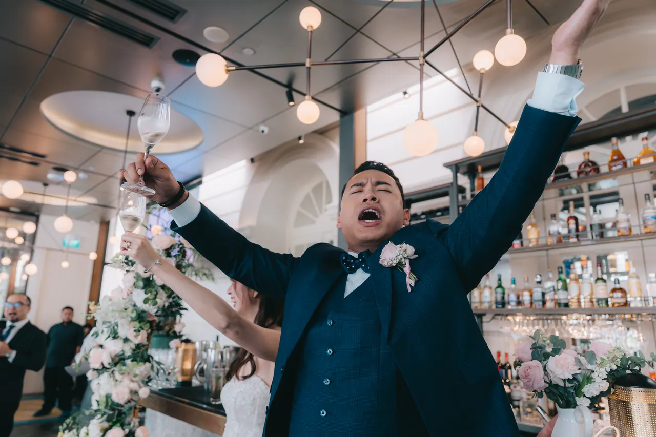 The groom throws both arms up mid-toast.