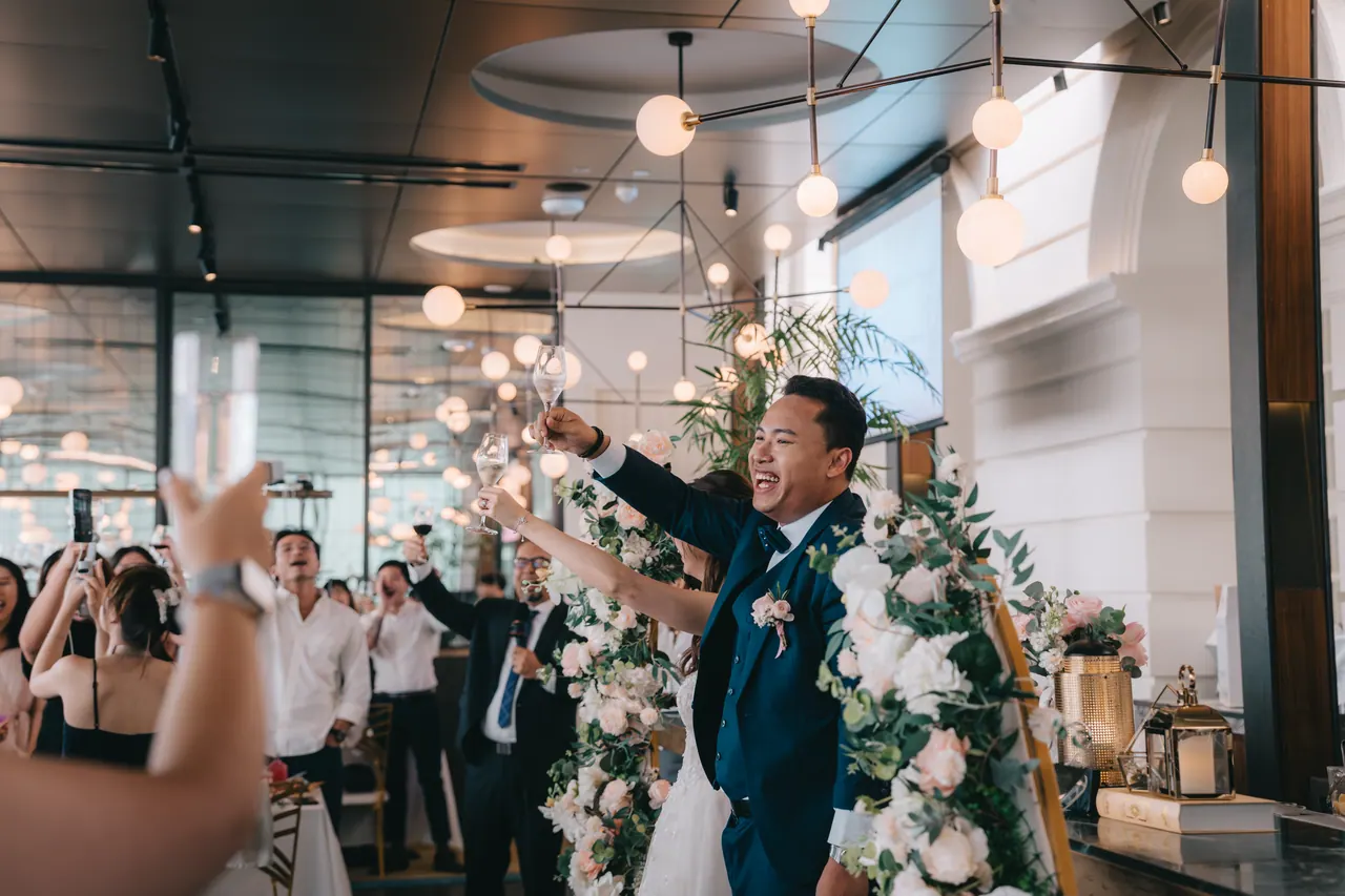 The room on its feet for the toast, the groom at its centre.