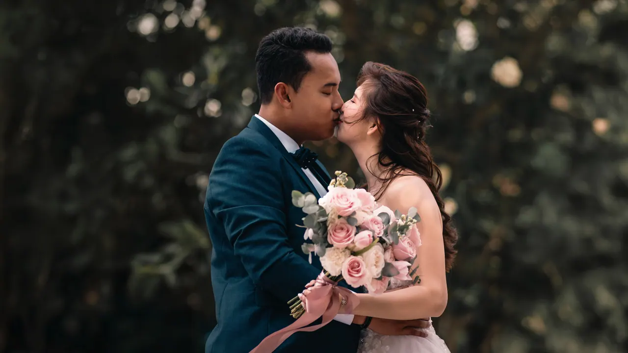 Bride and groom kiss in front of dark leaves, a bouquet of pink roses between them.
