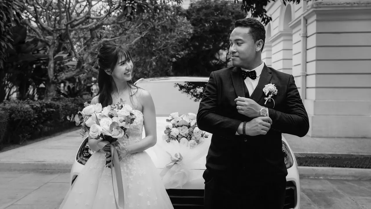 Black and white: the groom adjusts his cuff beside the bride, a decorated car behind them.