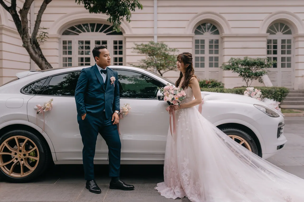 Bride and groom beside a white car in front of a building with arched windows.