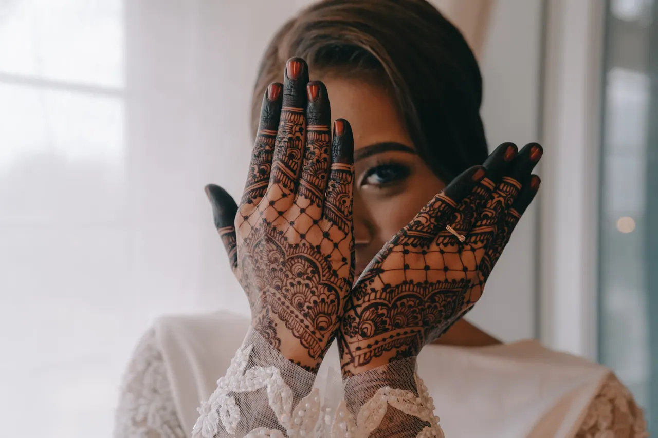 The bride raises her hennaed hands to her face, one eye showing between her fingers.