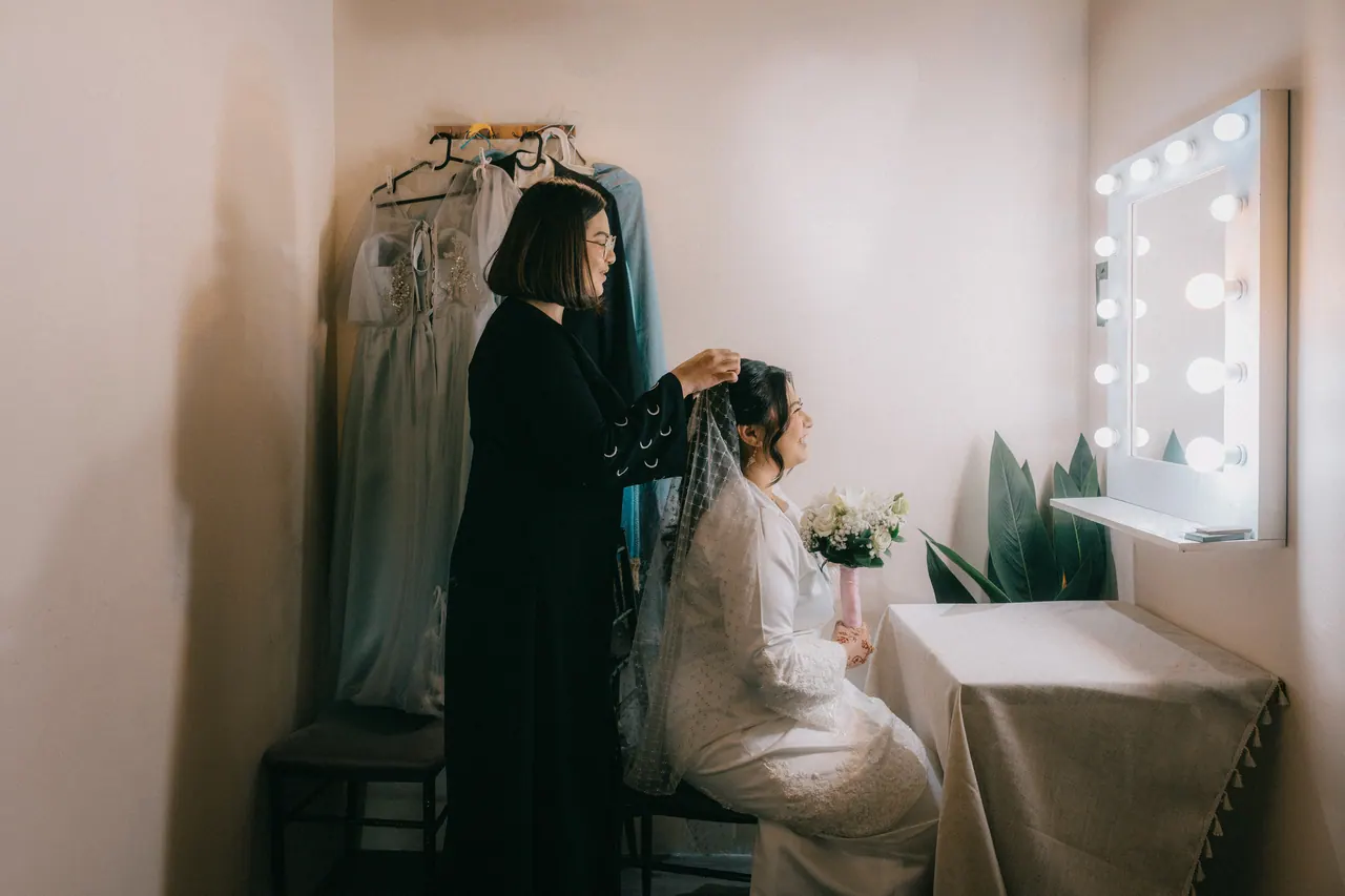 A woman in black arranges the bride’s veil at a dressing table.
