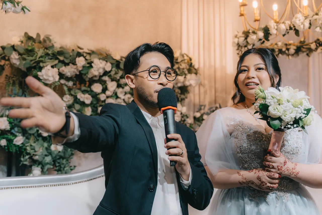 The groom gestures with a microphone mid-speech; the bride laughs.