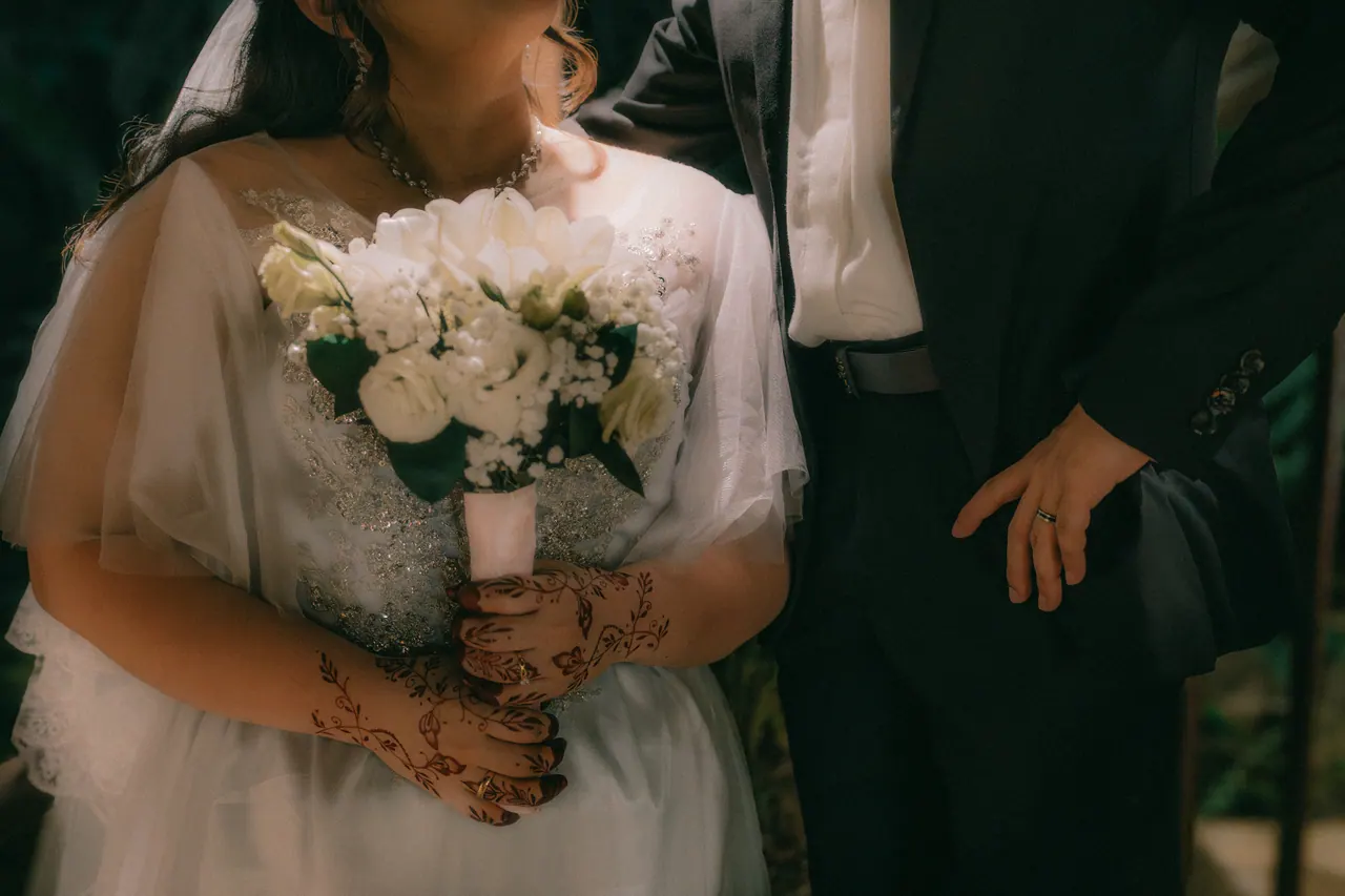 The bride’s hennaed hands around a white bouquet; the groom’s hand beside it.