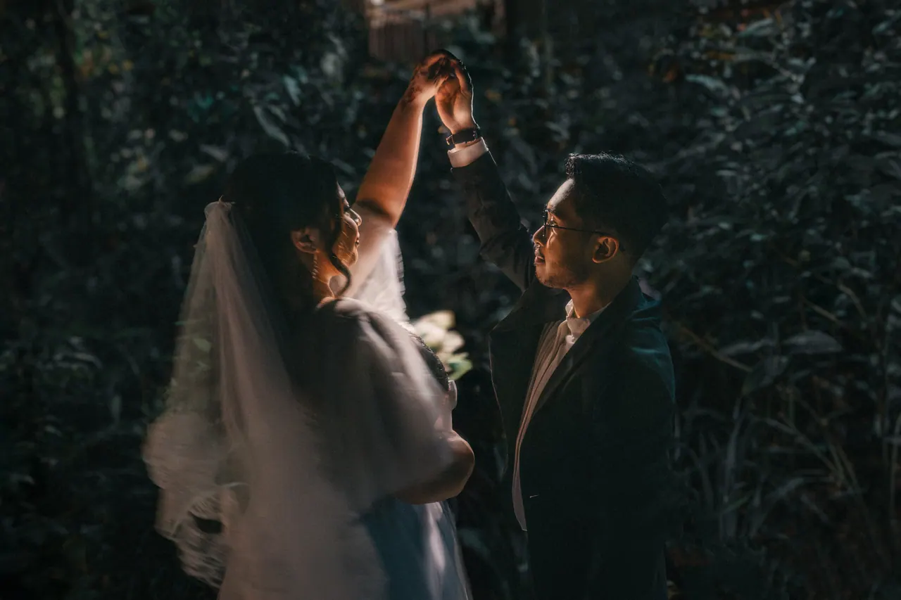 In a dark garden the groom spins the bride, her veil swinging.