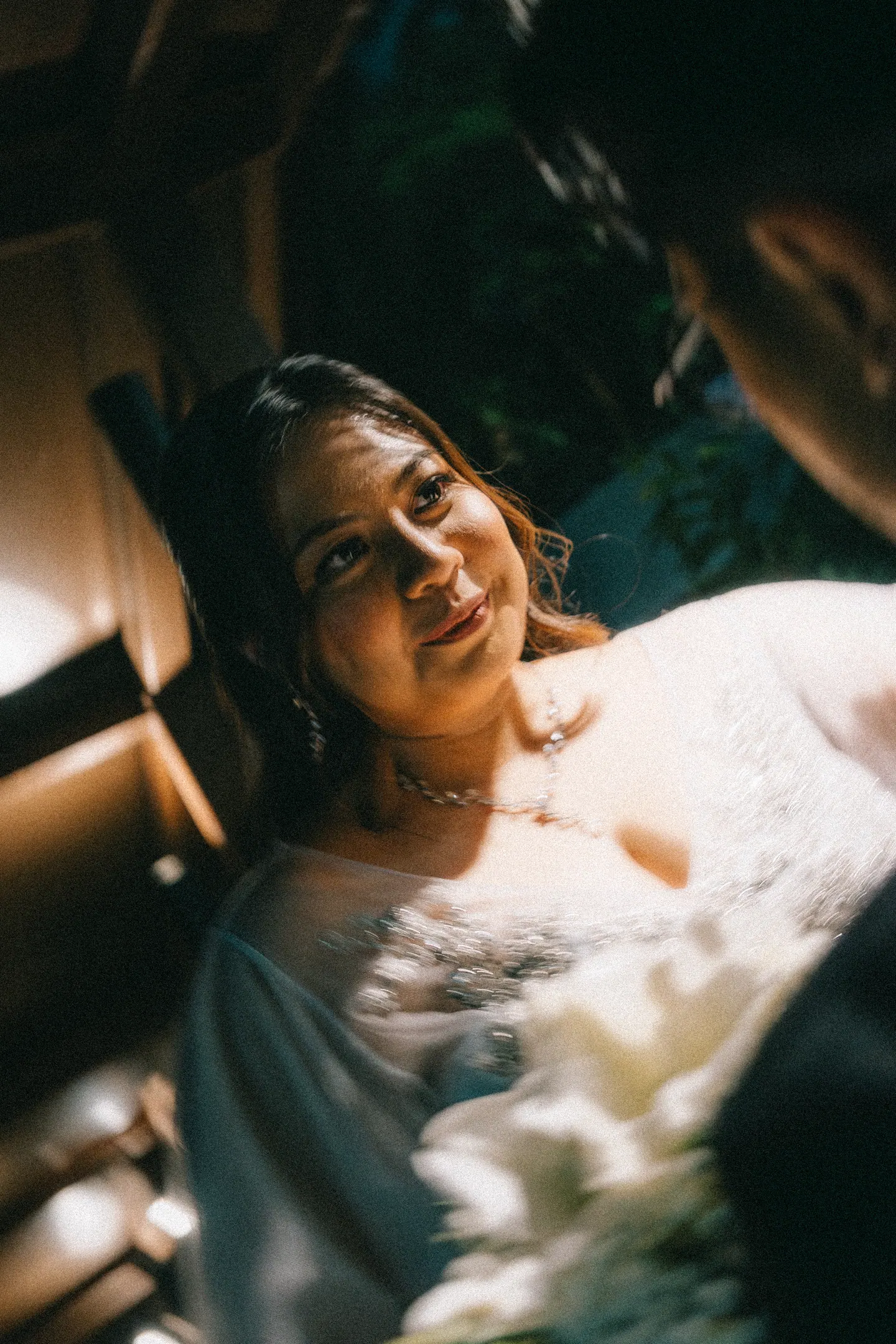 The bride looks up, lit by a warm lamp, flowers at her chest.