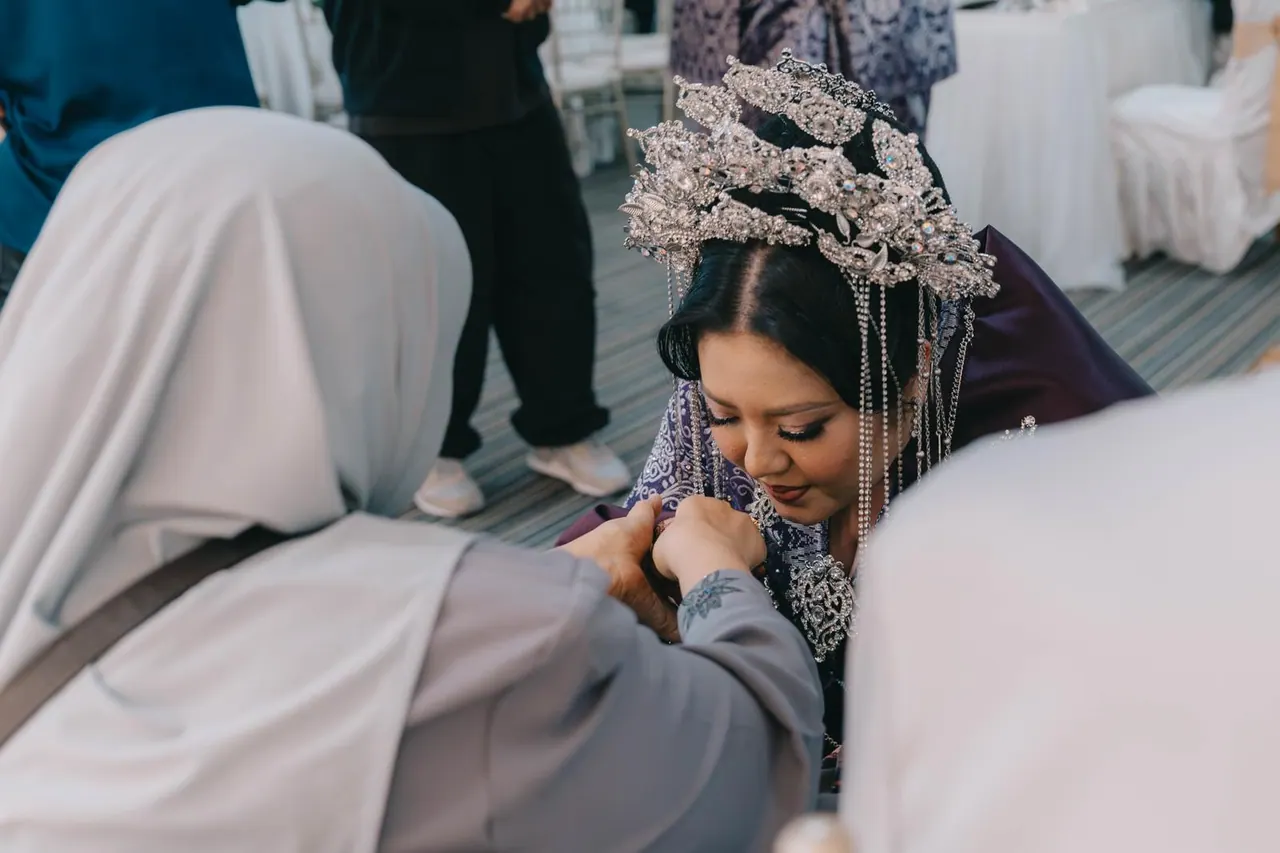 The bride, in purple with a tall silver headpiece, bows to kiss the hand of a guest in a grey headscarf.
