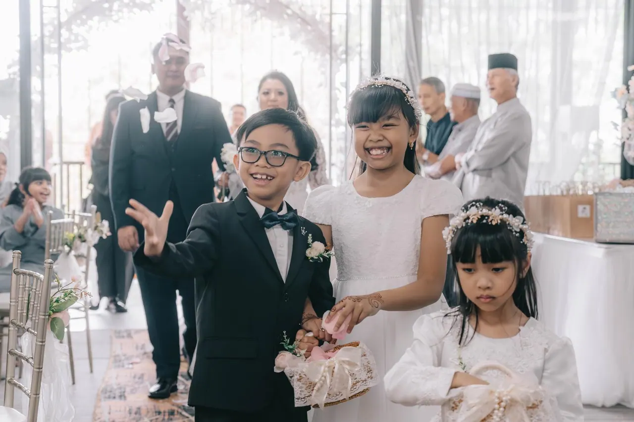 A boy in a small suit and a girl in white lead the way in, guests smiling behind them.