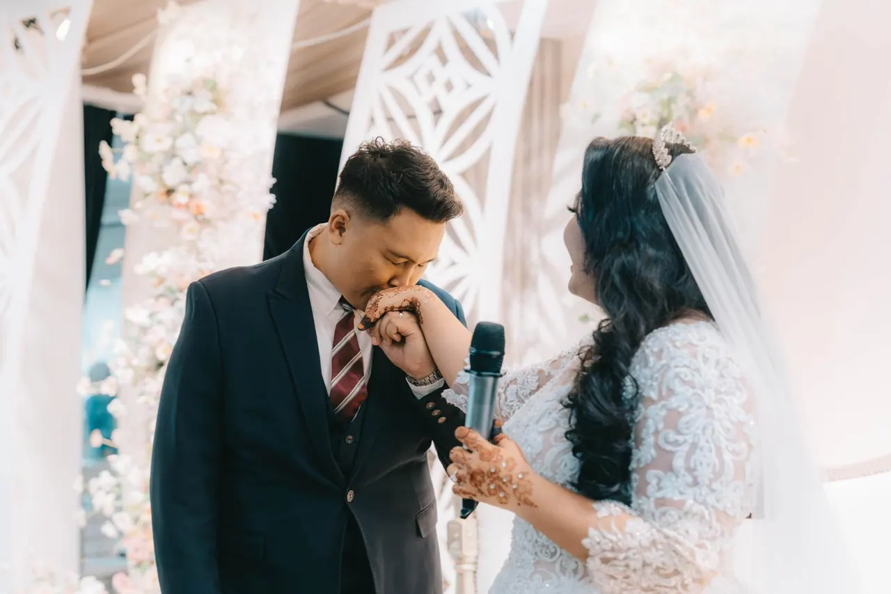 The groom, microphone in hand, kisses the bride's hand beneath a white floral arch.