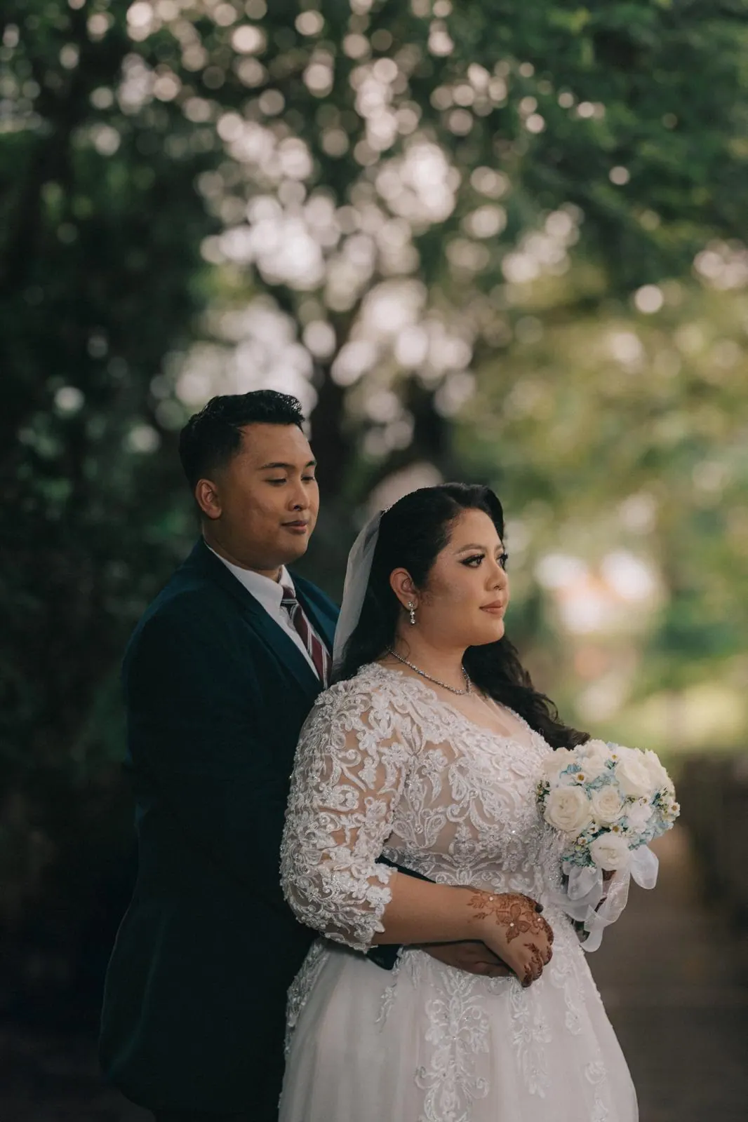 Bride and groom stand close among trees, she in a lace gown holding white roses.