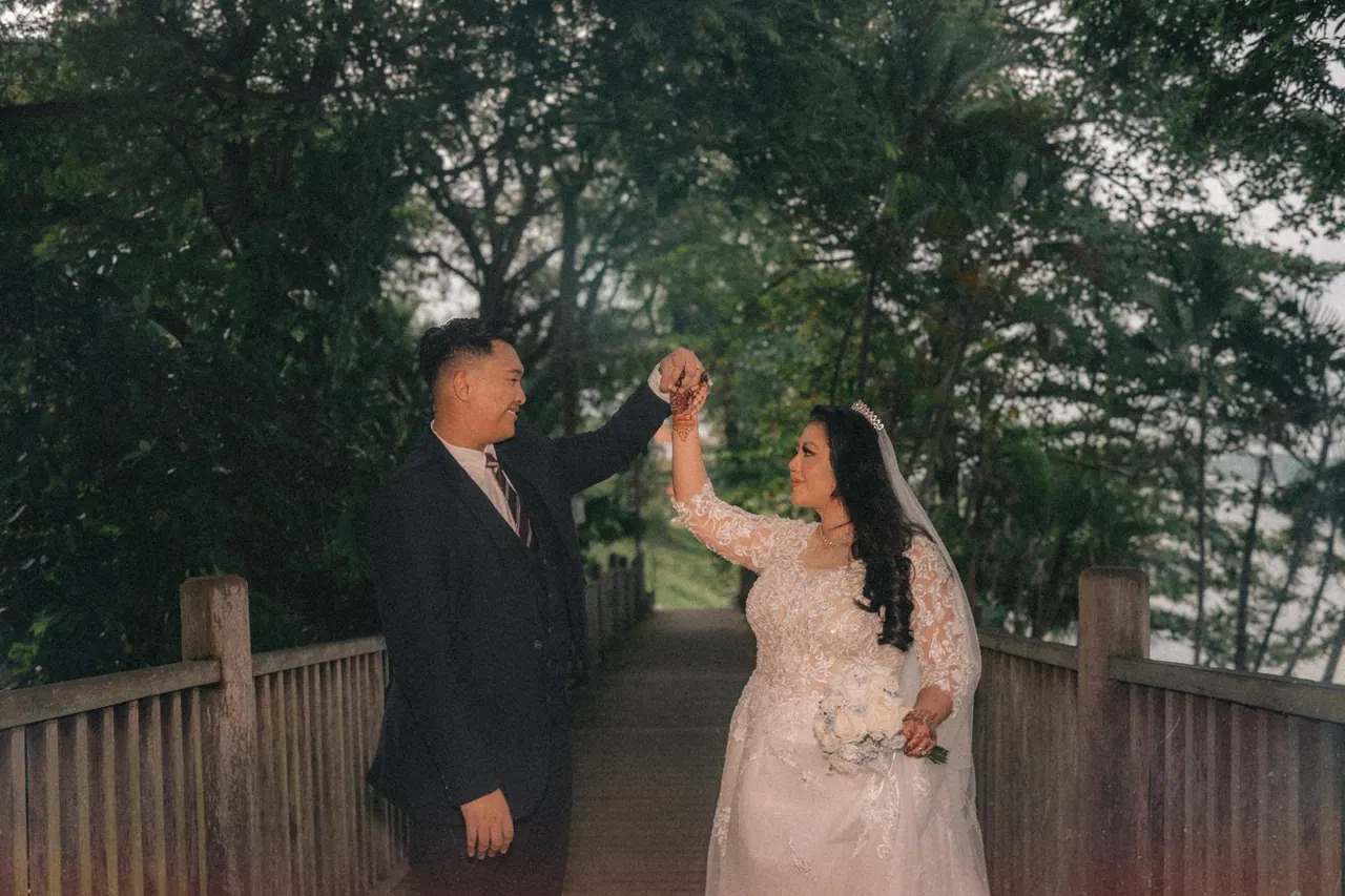 On a wooden boardwalk under trees, the groom lifts the bride's hand as she turns.