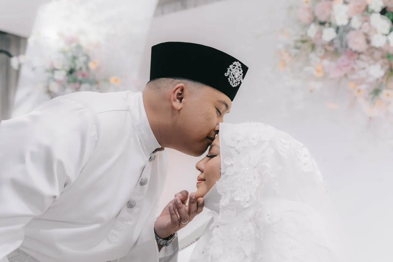 The groom in white kisses the bride's forehead as she touches his cheek.