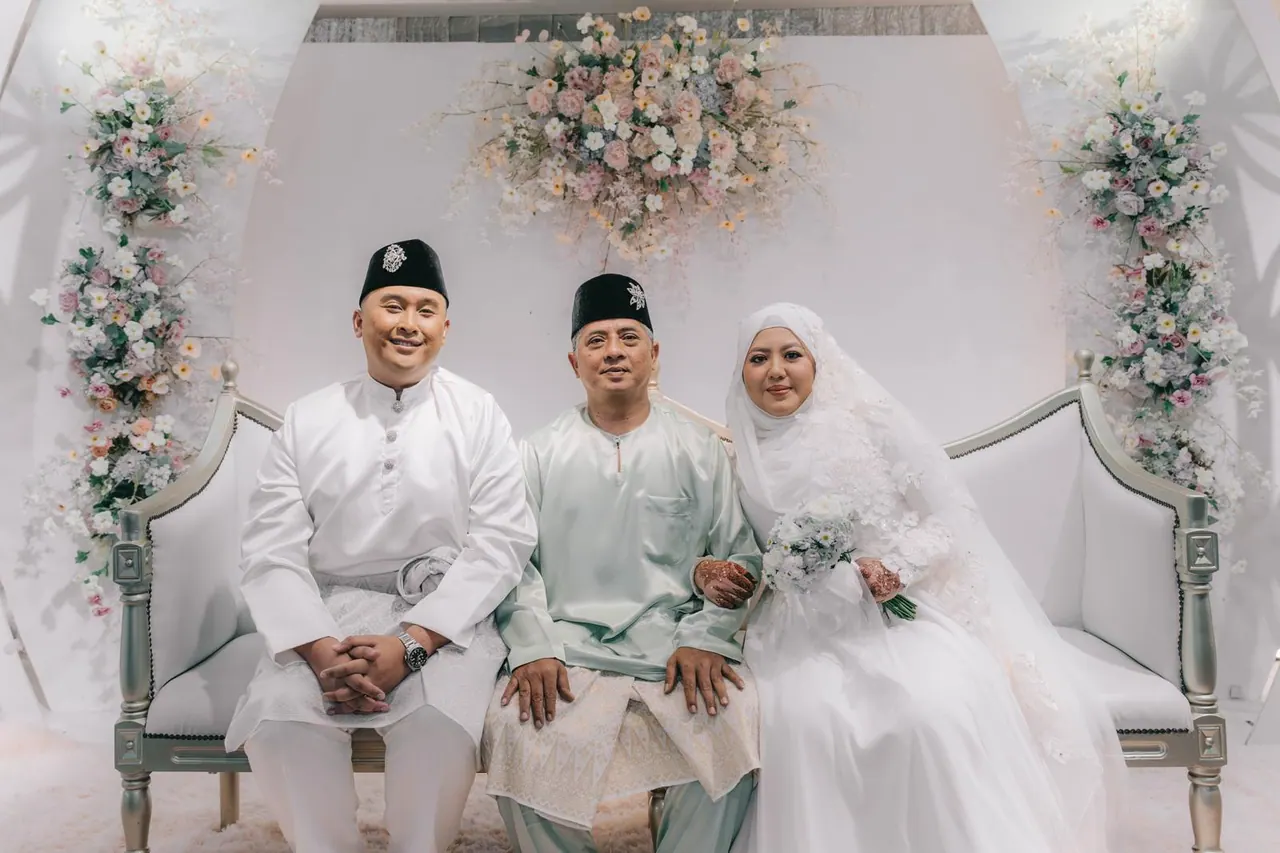 The groom, an older man in mint green and the bride sit together on a floral dais.