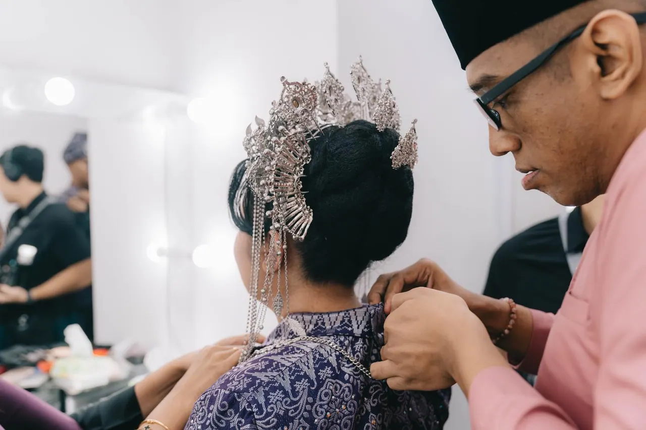 A man adjusts the back of the bride's silver headpiece in front of a dressing-room mirror.