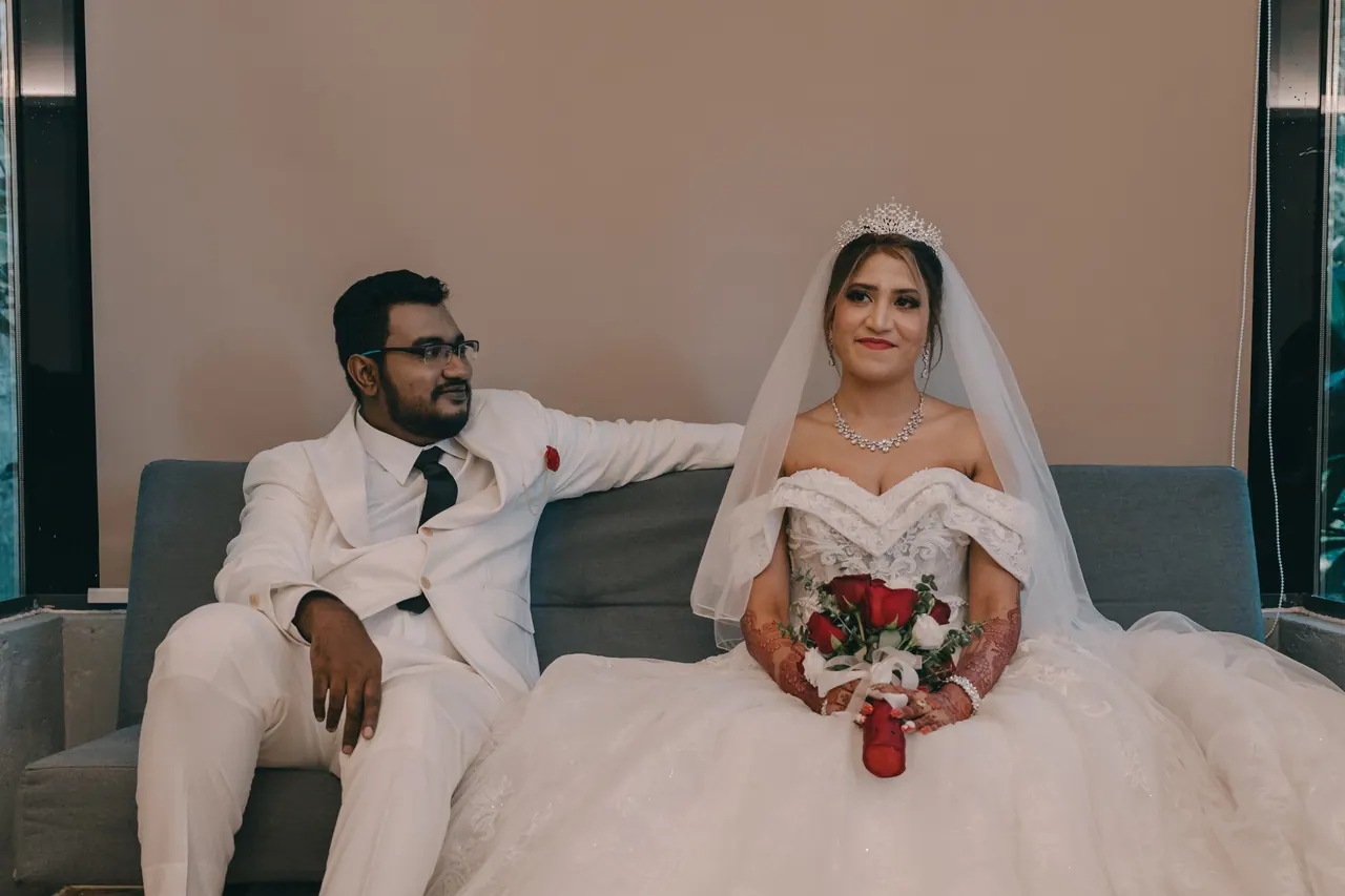 The couple sit on a sofa in white, she with red roses.