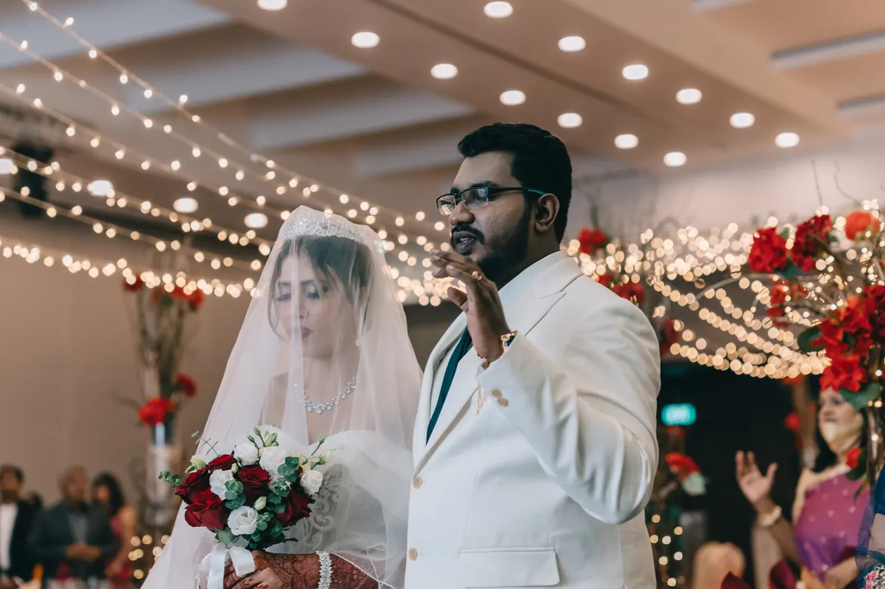 The couple stand under string lights, the groom’s hand raised.