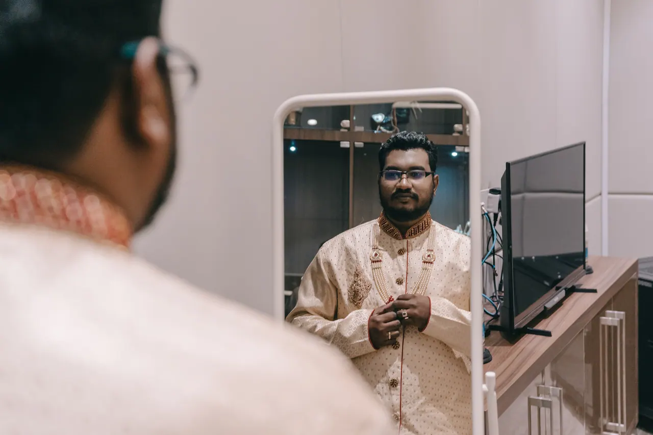The groom in a cream sherwani, seen in a mirror.