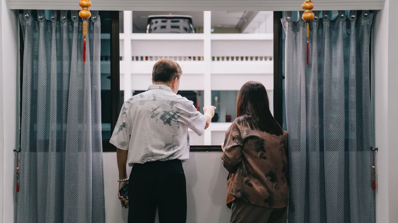 Two people stand at a window between grey curtains, looking out.