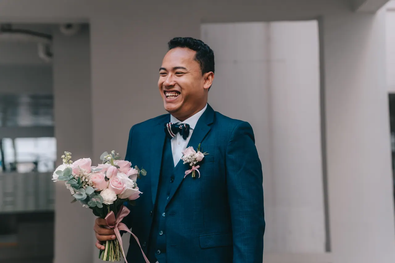 The groom laughs outside, a bouquet of pink roses in his hand.