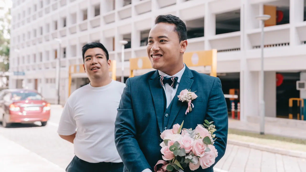 The groom smiles with a friend outside the flats, bouquet in hand.