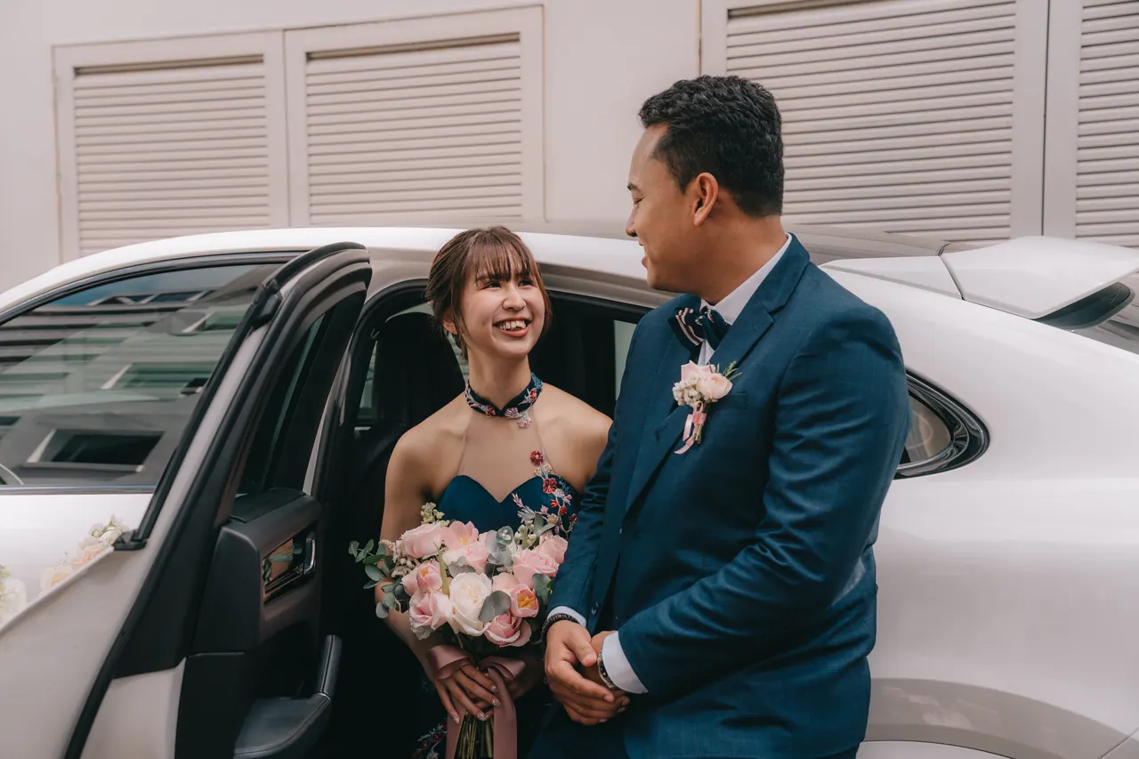 The couple stand by the open door of a white car.