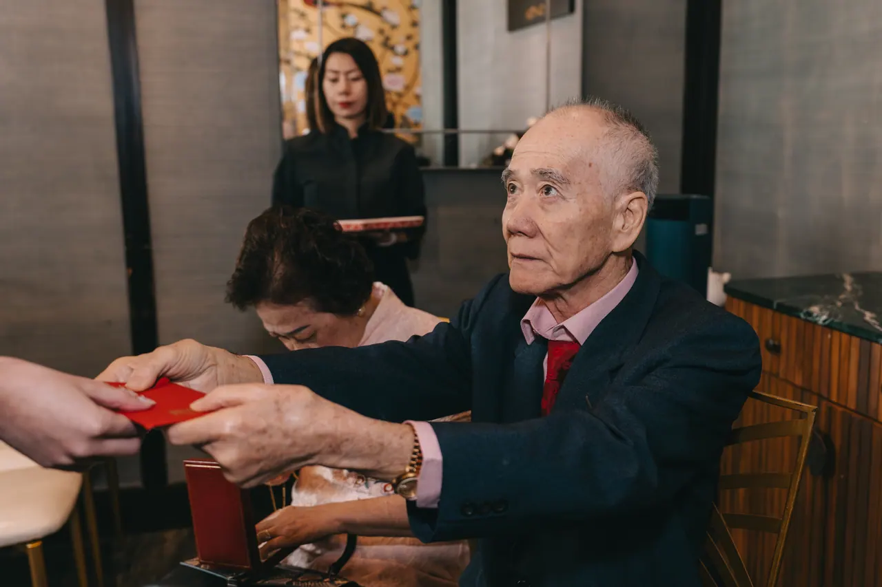An older man hands over a red packet across a table.
