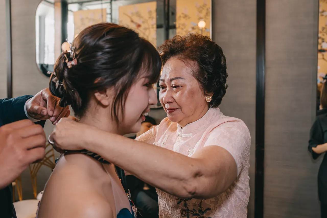 An older woman fastens a necklace on the bride, both close to tears.