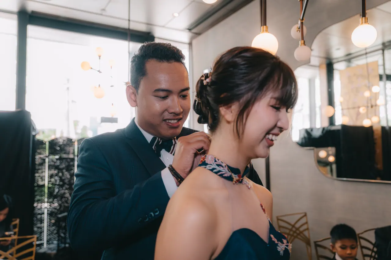 The groom fastens a necklace on the bride, who laughs.
