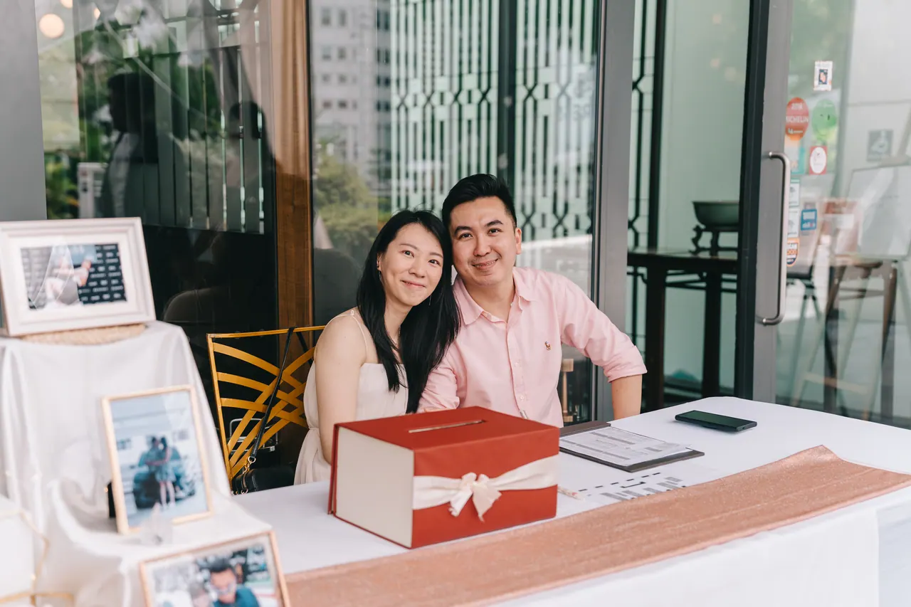 Two guests smile at the welcome table beside a red gift box.